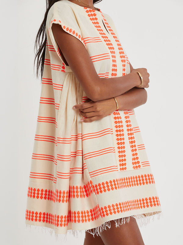 Woman Standing wearing lemlem handwoven ELINA Caftan featuring intricate electric orange toned Tibeb pattern on a cream biscuit background.