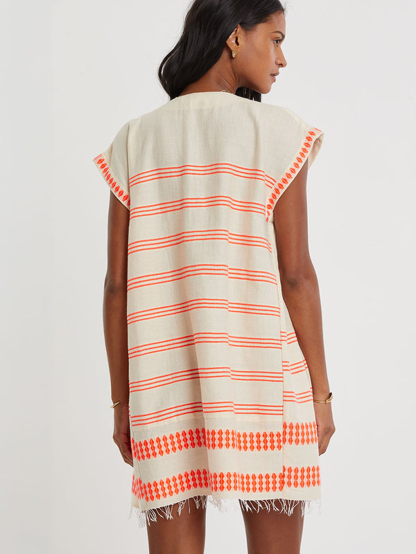Woman Standing wearing lemlem handwoven ELINA Caftan featuring intricate electric orange toned Tibeb pattern on a cream biscuit background.