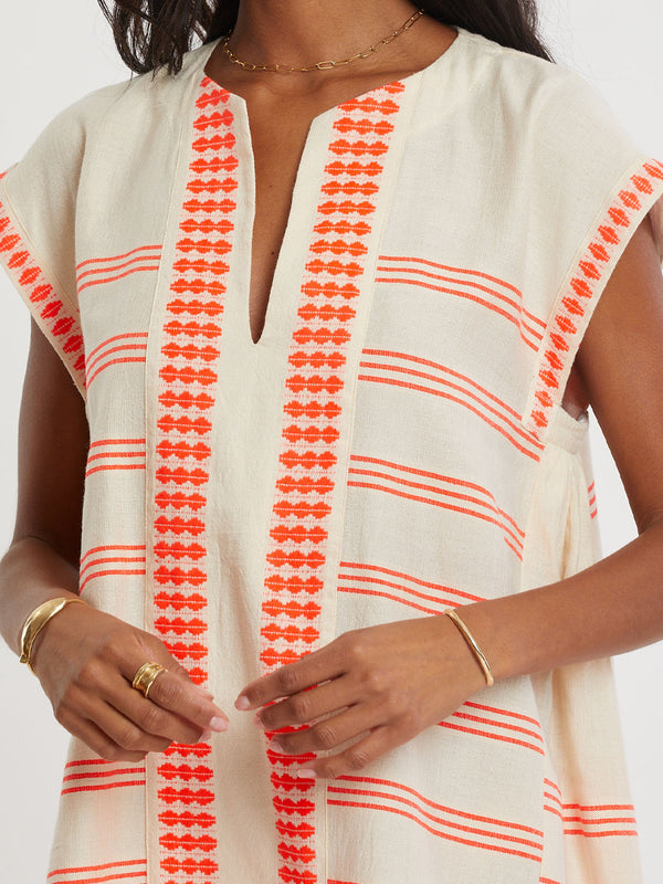 Woman Standing wearing lemlem handwoven ELINA Caftan featuring intricate electric orange toned Tibeb pattern on a cream biscuit background.