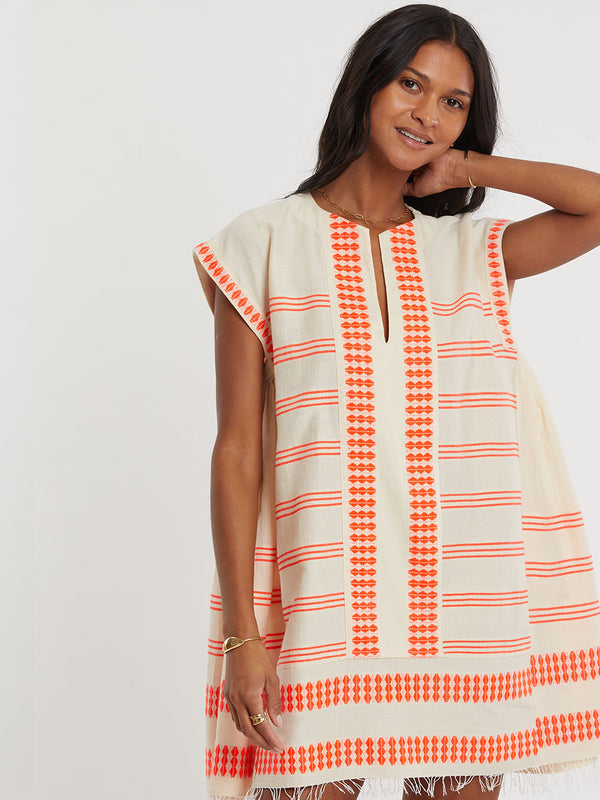 Woman Standing wearing lemlem handwoven ELINA Caftan featuring intricate electric orange toned Tibeb pattern on a cream biscuit background.