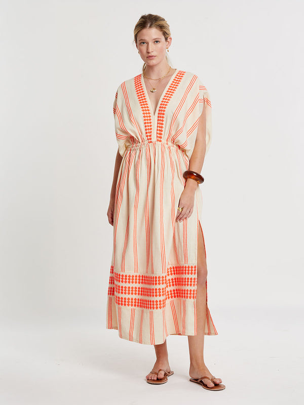 Woman Standing wearing lemlem handwoven LEILA Dress featuring intricate electric orange toned Tibeb pattern on a cream biscuit background.