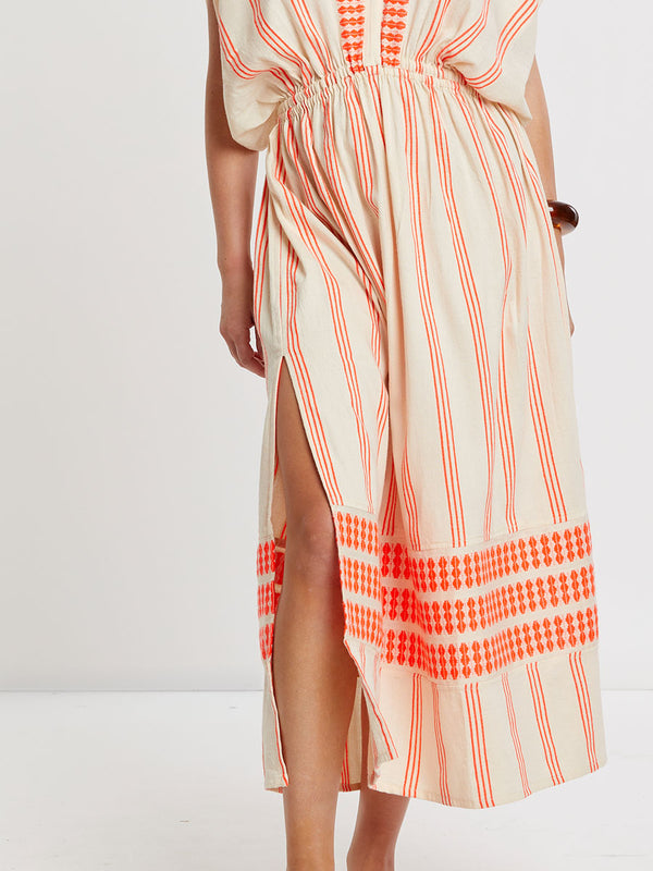 Woman Standing wearing lemlem handwoven LEILA Dress featuring intricate electric orange toned Tibeb pattern on a cream biscuit background.