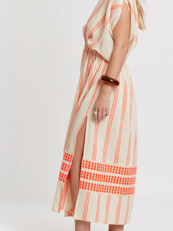 Woman Standing wearing lemlem handwoven LEILA Dress featuring intricate electric orange toned Tibeb pattern on a cream biscuit background.