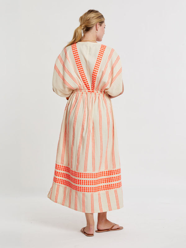 Woman Standing wearing lemlem handwoven LEILA Dress featuring intricate electric orange toned Tibeb pattern on a cream biscuit background.