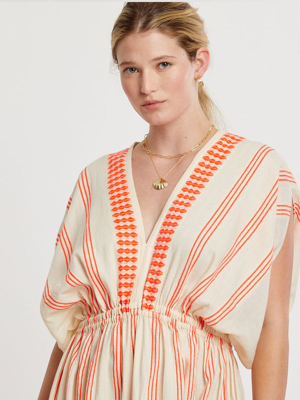 Woman Standing wearing lemlem handwoven LEILA Dress featuring intricate electric orange toned Tibeb pattern on a cream biscuit background.