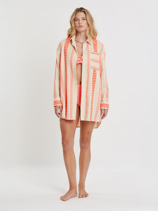 Woman Standing wearing lemlem handwoven MARIAM Shirt featuring intricate electric orange toned Tibeb pattern on a cream biscuit background.
