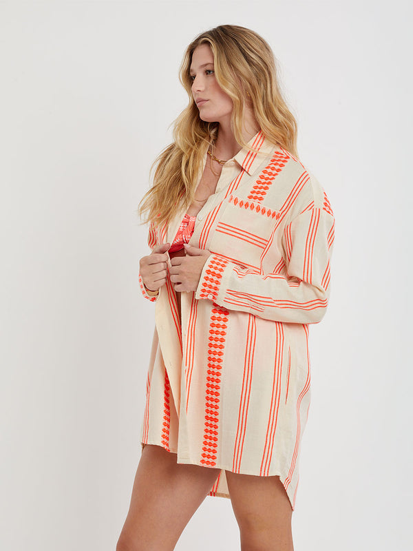 Woman Standing wearing lemlem handwoven MARIAM Shirt featuring intricate electric orange toned Tibeb pattern on a cream biscuit background.