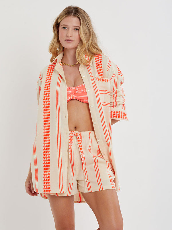 Woman Standing wearing lemlem handwoven MARIAM Shirt featuring intricate electric orange toned Tibeb pattern on a cream biscuit background.