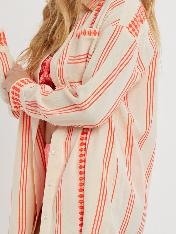 Woman Standing wearing lemlem handwoven MARIAM Shirt featuring intricate electric orange toned Tibeb pattern on a cream biscuit background.