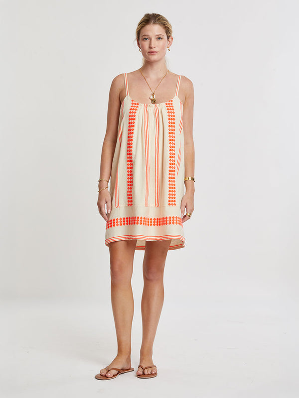 Woman Standing wearing lemlem handwoven ZINA Swing Dress featuring intricate electric orange toned Tibeb pattern on a cream biscuit background.