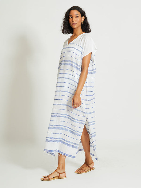 Side Shot of a Woman Standing Wearing lemlem DALILA | V Neck Caftan featuring crisp white fabric woven with diagonal dotted stripes in bright blue and a subtle textured weave