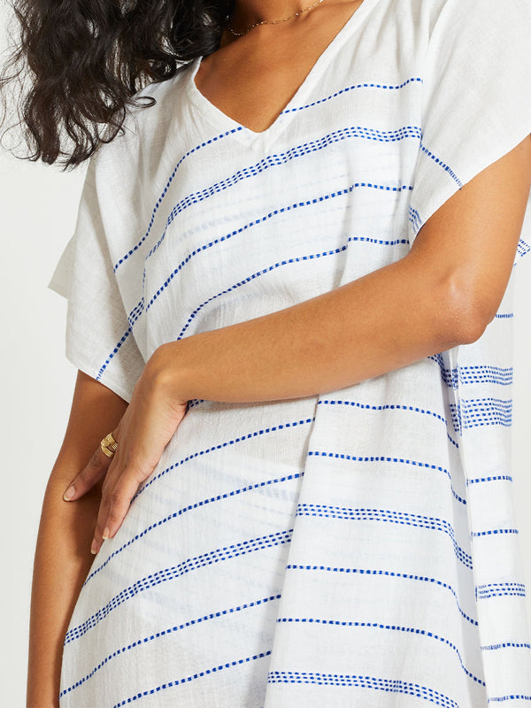 Close up on a Woman Standing Wearing lemlem DALILA | V Neck Caftan featuring crisp white fabric woven with diagonal dotted stripes in bright blue and a subtle textured weave