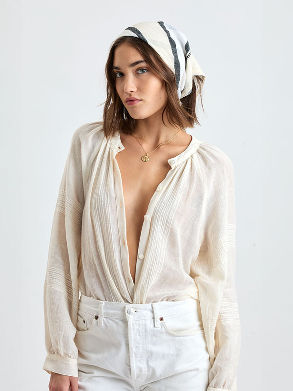 Woman standing wearing lemlem handwoven DANA Bandana featuring  refined palette of crisp white, soft cream, and deep black and white jeans along with cream handwoven MITA Blouse