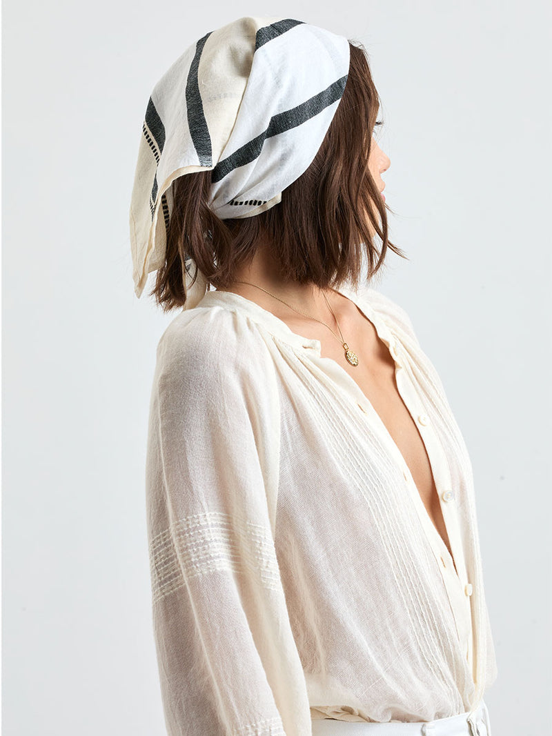 Woman standing wearing lemlem handwoven DANA Bandana featuring  refined palette of crisp white, soft cream, and deep black and white jeans along with cream handwoven MITA Blouse