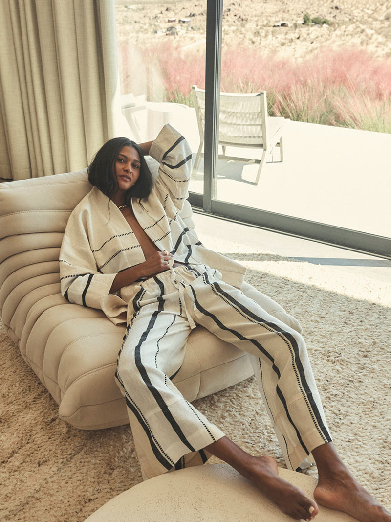 Woman sitting on a banch wearing lemlem handwoven jacket and pants featuring black stripe pattern on cream background