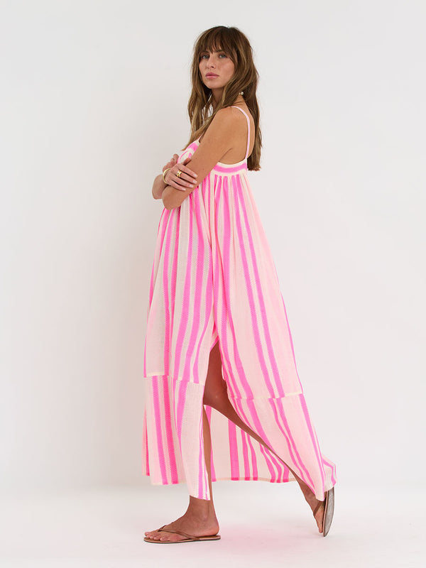 Woman Standing wearing lemlem handwoven EDA Slip Dress featuring a ribbon stripe pattern with a picot edge in vibrant neon pink on a soft vanilla background 