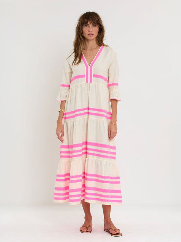 Woman Standing wearing lemlem handwoven HAWI Flutter Dress featuring a ribbon stripe pattern with a picot edge in vibrant neon pink on a soft vanilla background.