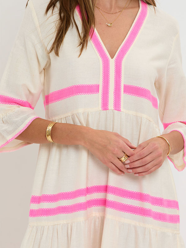 Woman Standing wearing lemlem handwoven HAWI Flutter Dress featuring a ribbon stripe pattern with a picot edge in vibrant neon pink on a soft vanilla background.