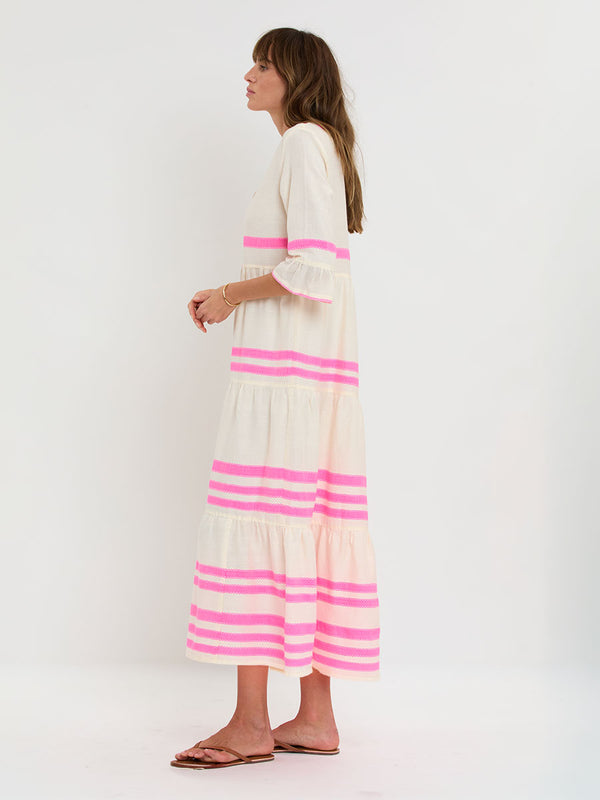 Woman Standing wearing lemlem handwoven HANNA Flutter Dress featuring a ribbon stripe pattern with a picot edge in vibrant neon pink on a soft vanilla background.
