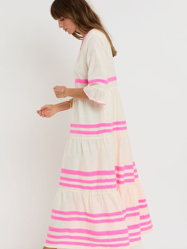 Woman Standing wearing lemlem handwoven HAWI Flutter Dress featuring a ribbon stripe pattern with a picot edge in vibrant neon pink on a soft vanilla background.