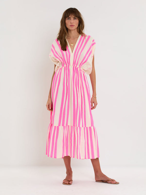 Woman Standing wearing lemlem handwoven LEILA Dress featuring a ribbon stripe pattern with a picot edge in vibrant neon pink on a soft vanilla background 