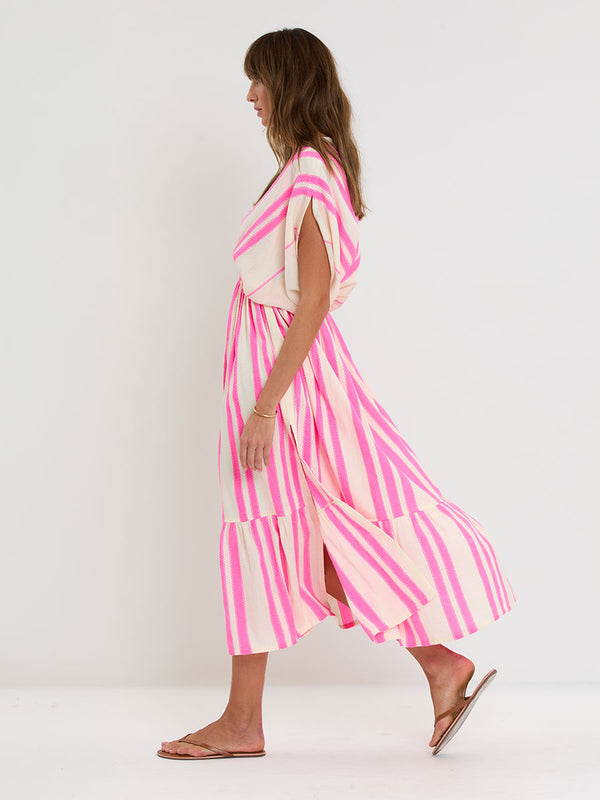 Woman Standing wearing lemlem handwoven LEILA Dress featuring a ribbon stripe pattern with a picot edge in vibrant neon pink on a soft vanilla background 