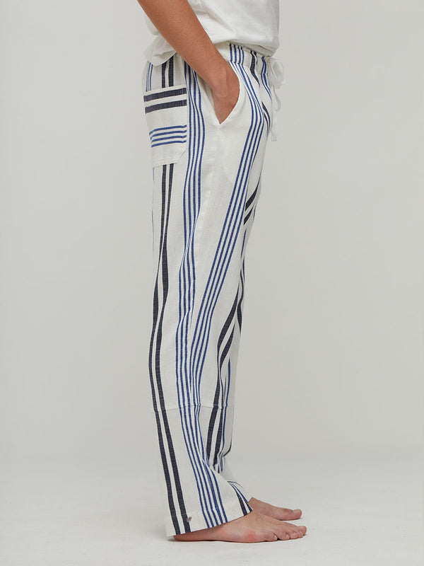 A side view of a person wearing lemlem handwoven white pants with vertical navy and blue stripes, paired with a white top, standing barefoot against a light gray studio background.