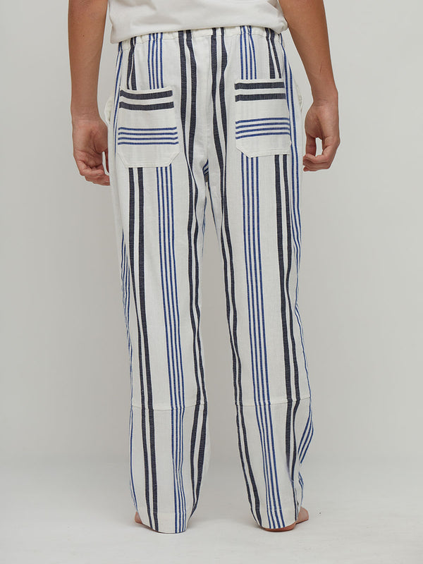 A person shown from the back wearing lemlem handwoven loose white pants with vertical navy and blue stripes and two back patch pockets, against a plain light gray studio background.