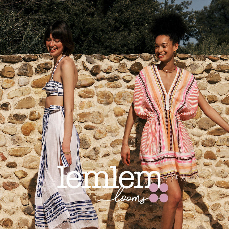 Two woman standing against an old stone wall with the lemlem looms logo overlayed. One woman is a white and blue bandeau bikini top and match wrap skirt. The other women is wearing a pink with brown graphic stripe dress. 