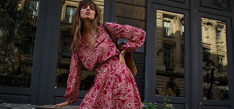 Woman in a red patterned dress standing in an urban setting with reflective windows.