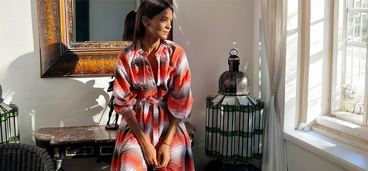 Liya Kebede sitting in an appartment by a window wearing a long handwoven lemlem dress with white, red and brown thin vertical stripes.