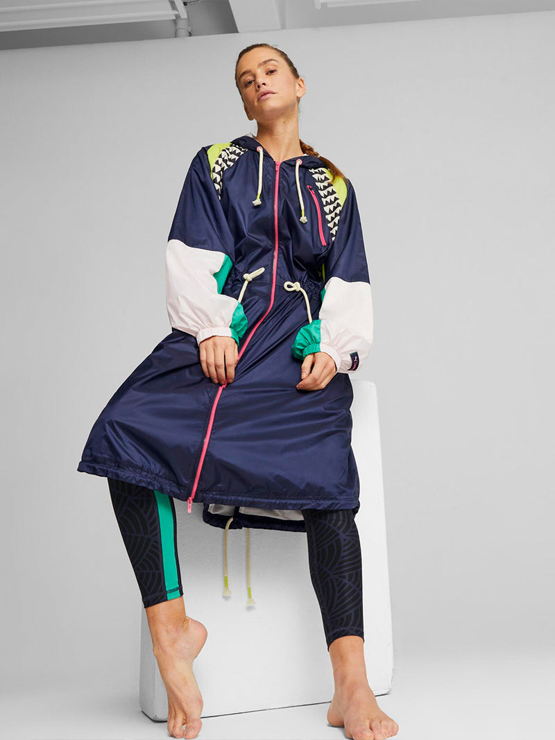 Woman sitting on a white seat wearing Puma x lemlem Anorak featuring Navy color, lemlem triangle pattern and color block details in green and yellow colors and Puma x lemlem High Waist 7/8 Leggings featuring navy color, scallop print and color block side stripes in Green and Pink Colors