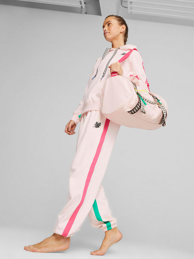 Woman walking wearing Puma x lemlem fleece oversized hoodie and joggers in Frosty Pink Color featuring color pop side stripes in pink and green colors and handsketched puma cat logo and Puma x lemlem studio bag in Frosty Pink Color