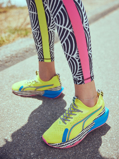 Close up on Woman Standing Wearing Puma x lemlem PWR XX NITRO™ LUXE sneakers featuring Yellow Burst, Racing Blue and Bright Pink colors and also featuring lemlem triangle pattern and high waist leggings featuring color block side stripes, scallop print in ghost pepper and black colors