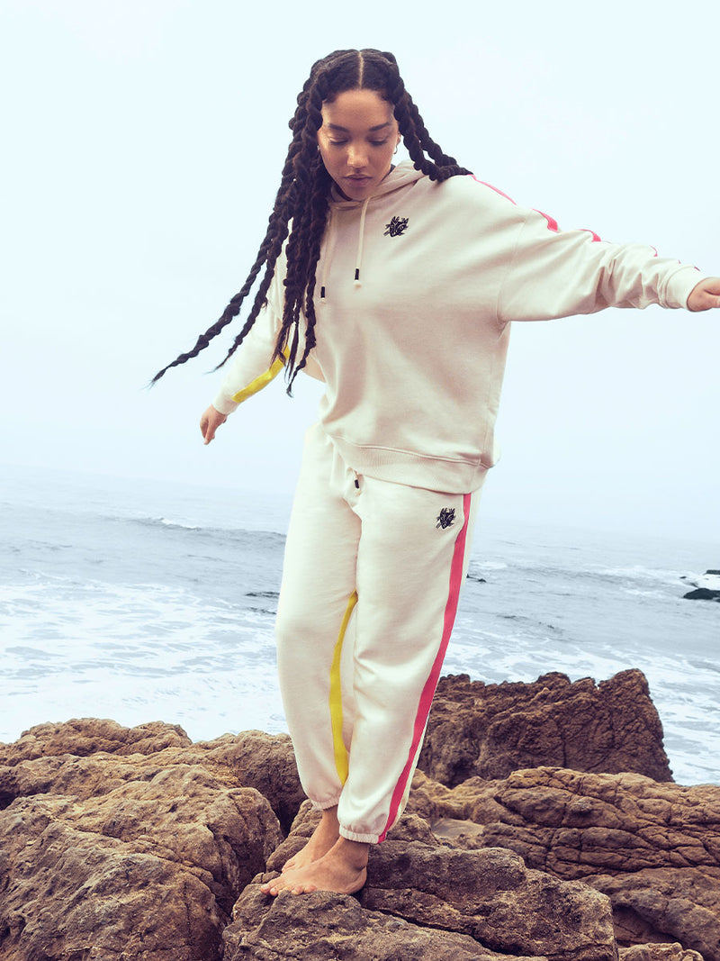 Woman standing on the beach wearing Puma x lemlem oversized hoodie and joggers in a Ghost Pepper Color featuring hand sketched cat logo and color block stripes in bright pink and yellow colors.