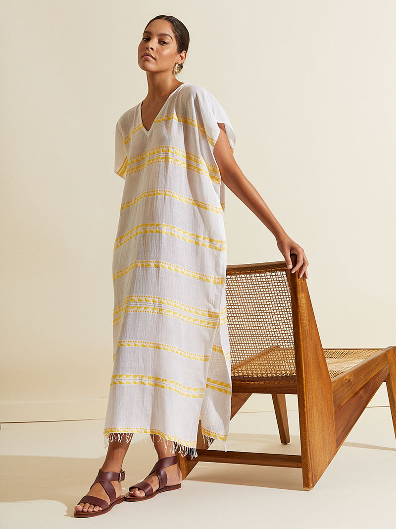 Woman standing by a chair wearing the Abeba Classic Caftan featuring the yellow signature Tibeb pattern and gold lurex highlights on a white background.