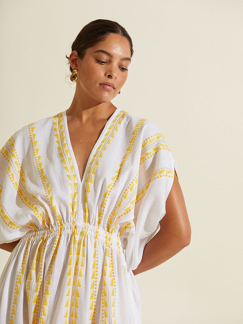 Woman standing with her arms behind her back wearing the Abeba Plunge Neck Dress featuring the yellow signature Tibeb pattern and gold lurex highlights on a white background.