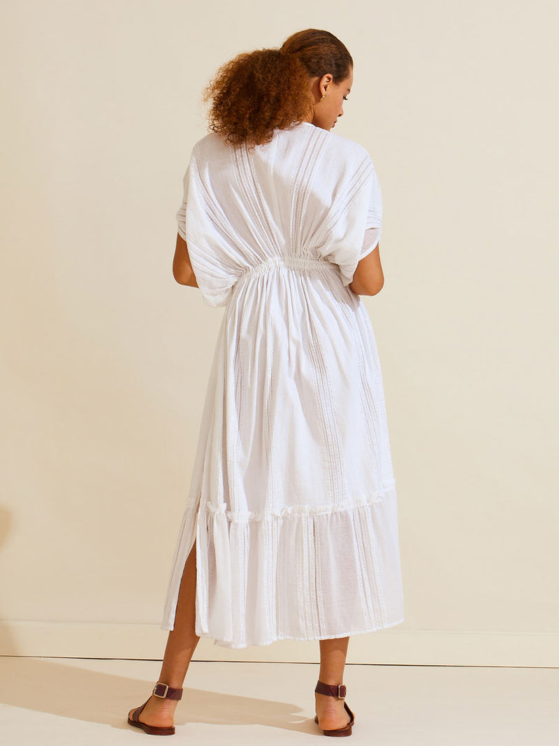 Back view of a woman standing wearing a white Abira plunge neck maxi dress with stitches of silver lurex
