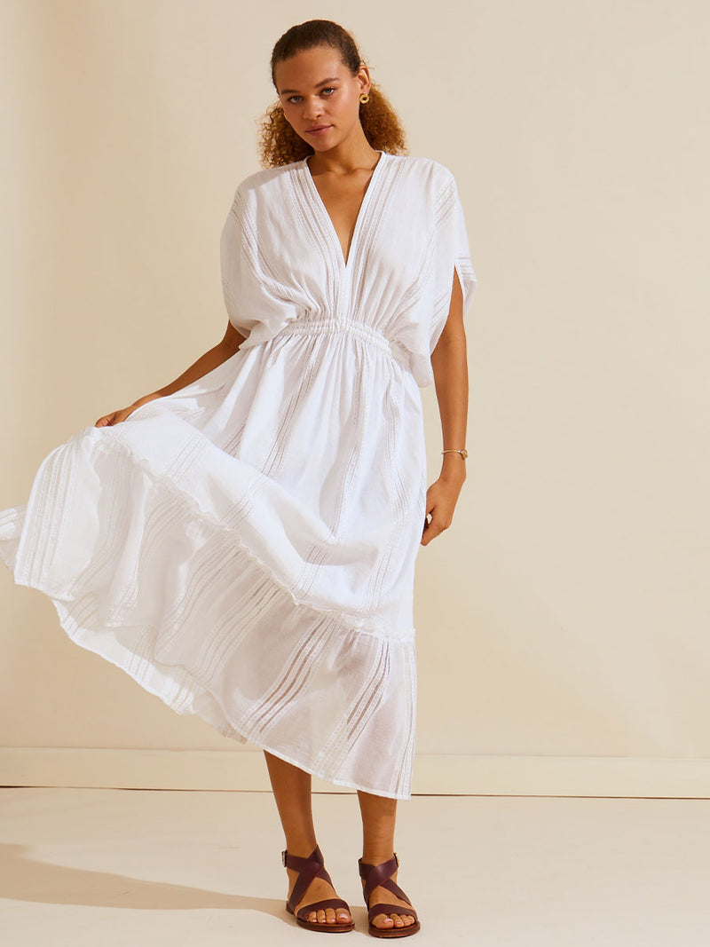 Woman standing wearing a white Abira plunge neck maxi dress with stitches of silver lurex