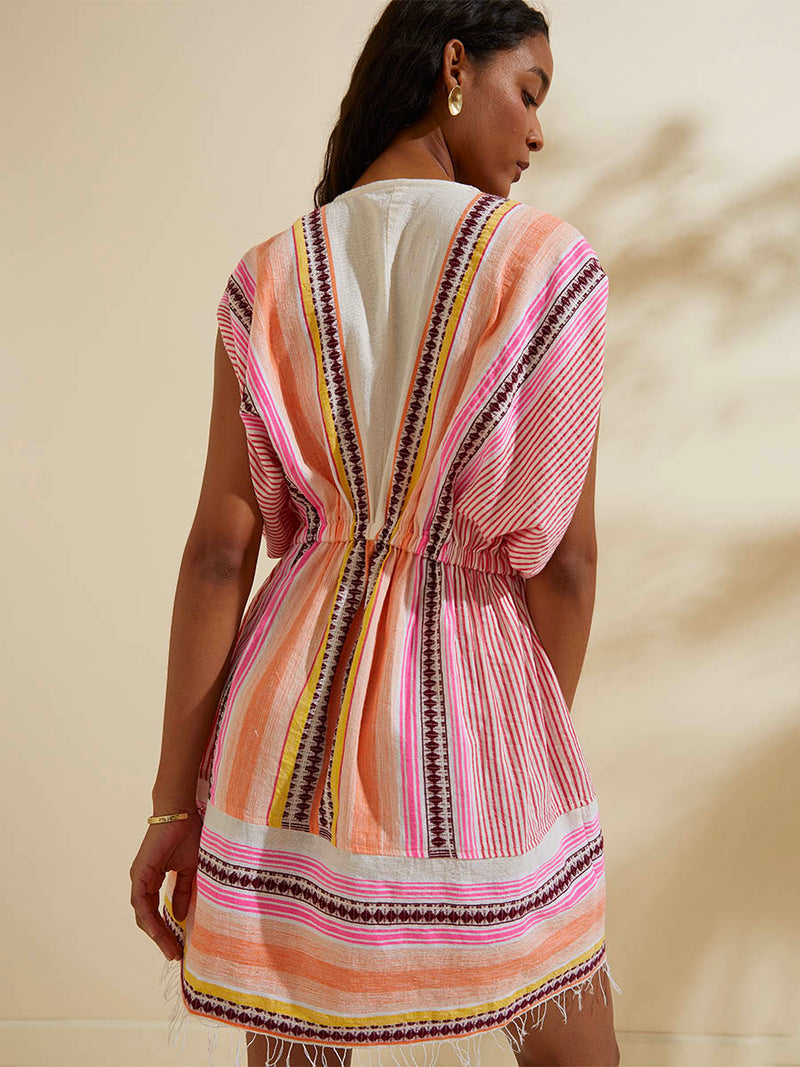 Back view of a woman standing  wearing the Cirq Short Plunge Neck Dress featuring pink, orange and yellow stripes and signature Tibeb design in burgundy bands.