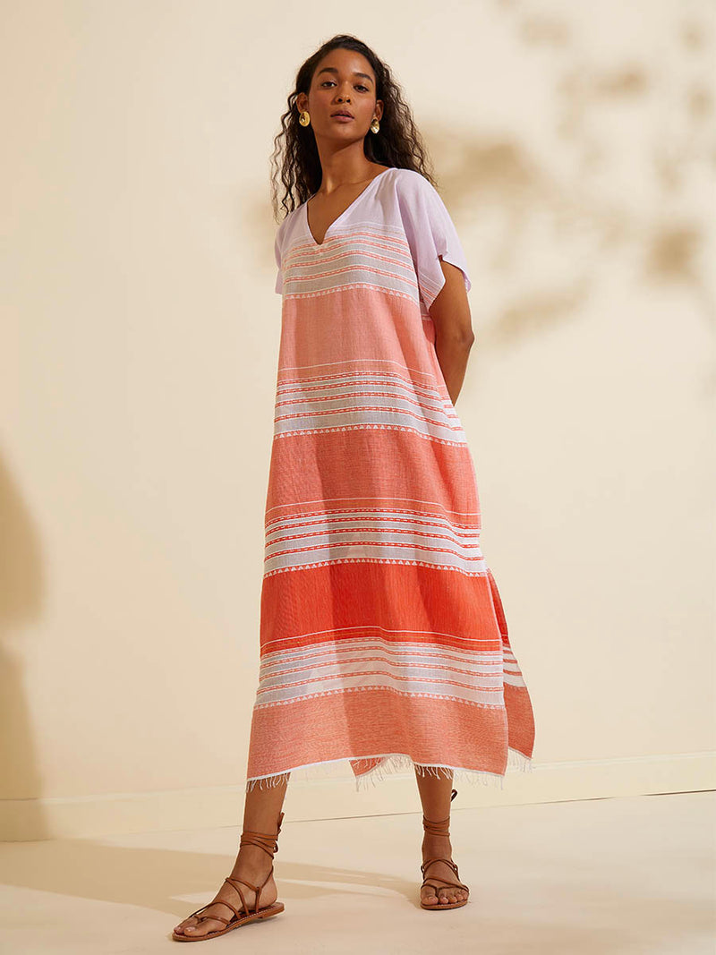 Woman standing wearing the Eshal Caftan Dress featuring white doted stripes with gradiant orange and tangerine bands on a lilac and white background.
