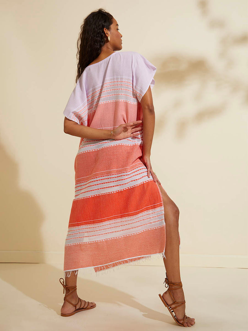 Back view of a woman standing wearing the Eshal Caftan Dress featuring white doted stripes with gradiant orange and tangerine bands on a lilac and white background.