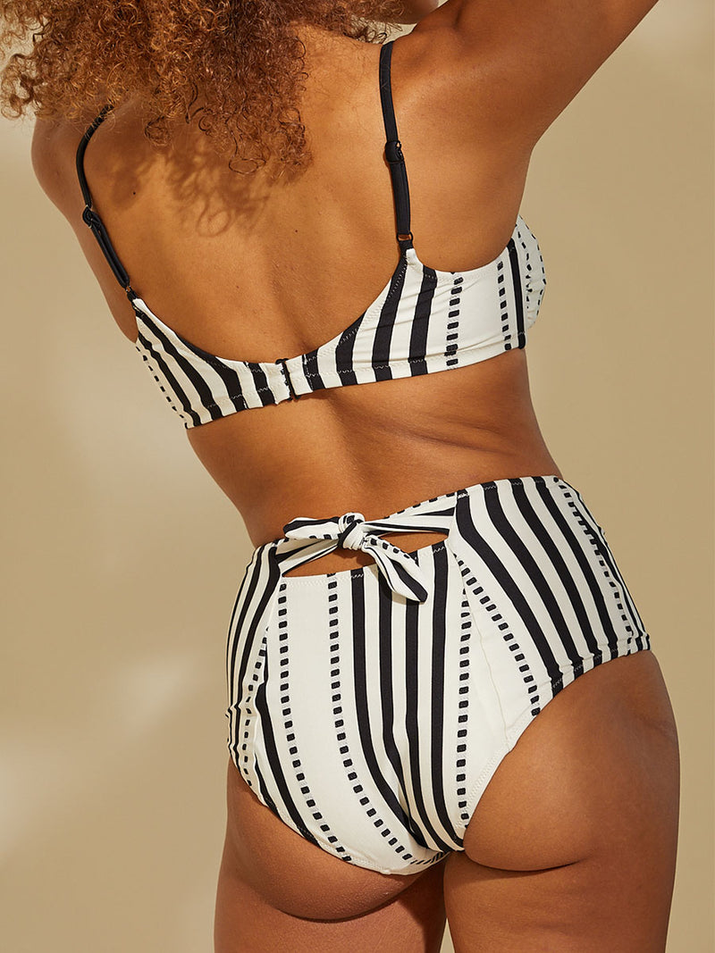 Back view of a woman standing with her arms up wearing the Eshe Bralette Top and matching high waist bikini bottom featuring architectural and textured black stripes and dotted lines on an off white background.