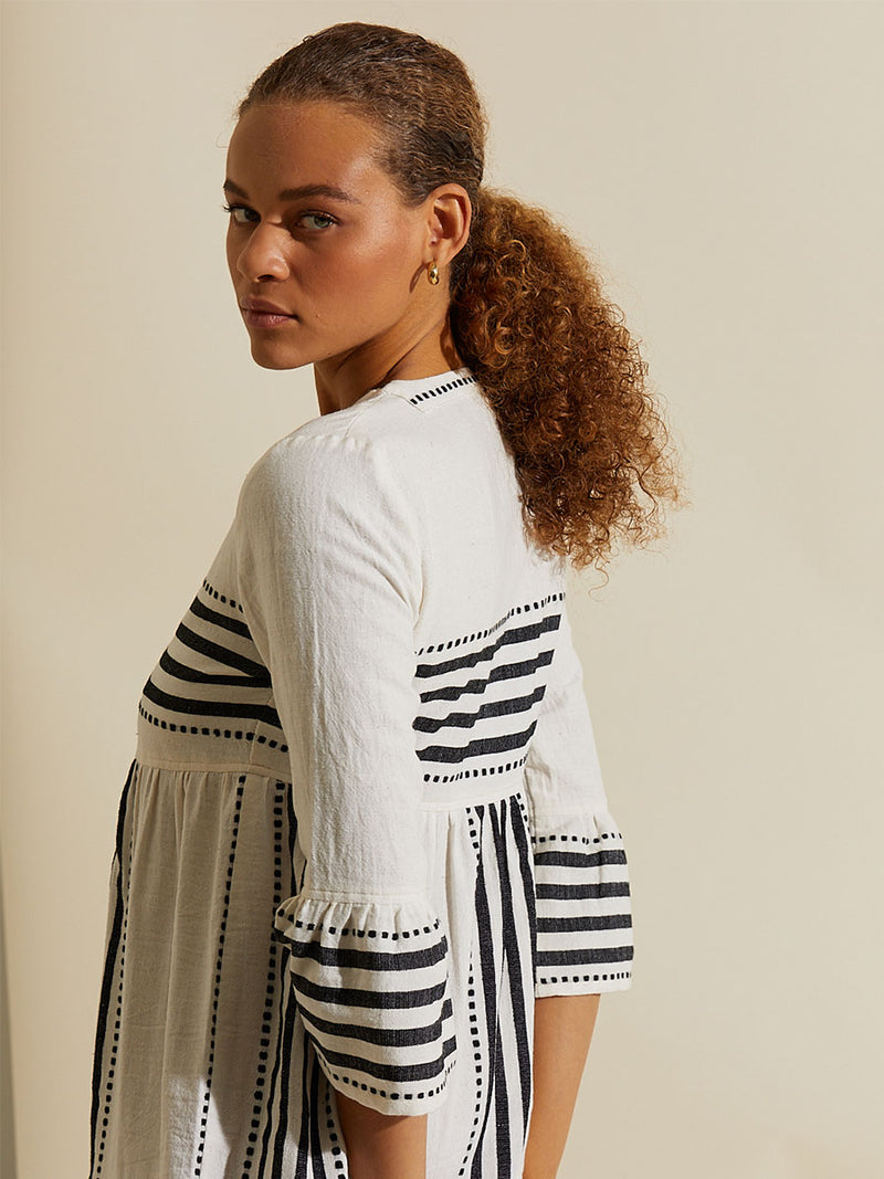 Side view of a woman looking over her shoulder wearing the Eshe Flutter Dress featuring architectural and textured black stripes and dotted lines on an off white background.