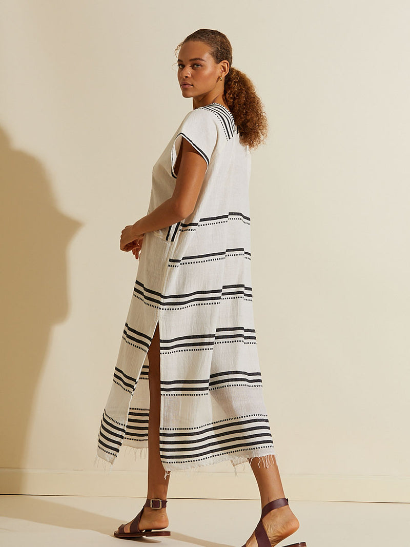 Side view of a woman walking wearing the Eshe Long Caftan Dress featuring architectural and textured black stripes and dotted lines on an off white background.