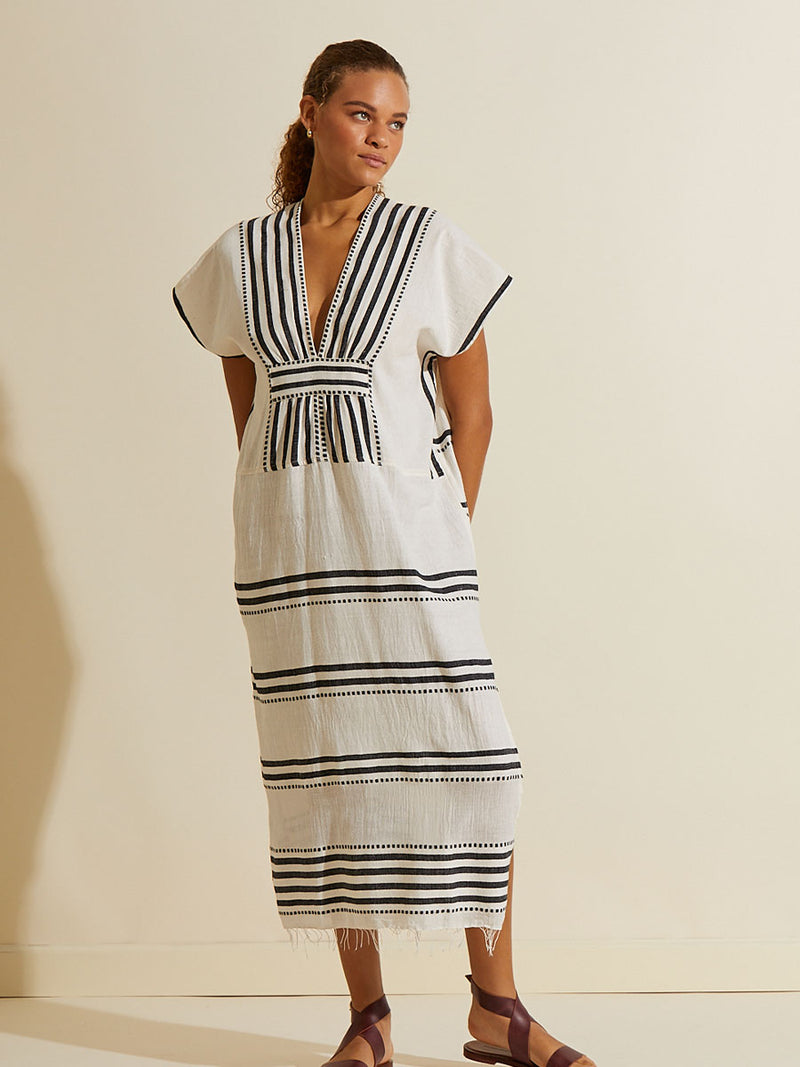 Woman standing with her hand in her back wearing the Eshe Long Caftan Dress featuring architectural and textured black stripes and dotted lines on an off white background.