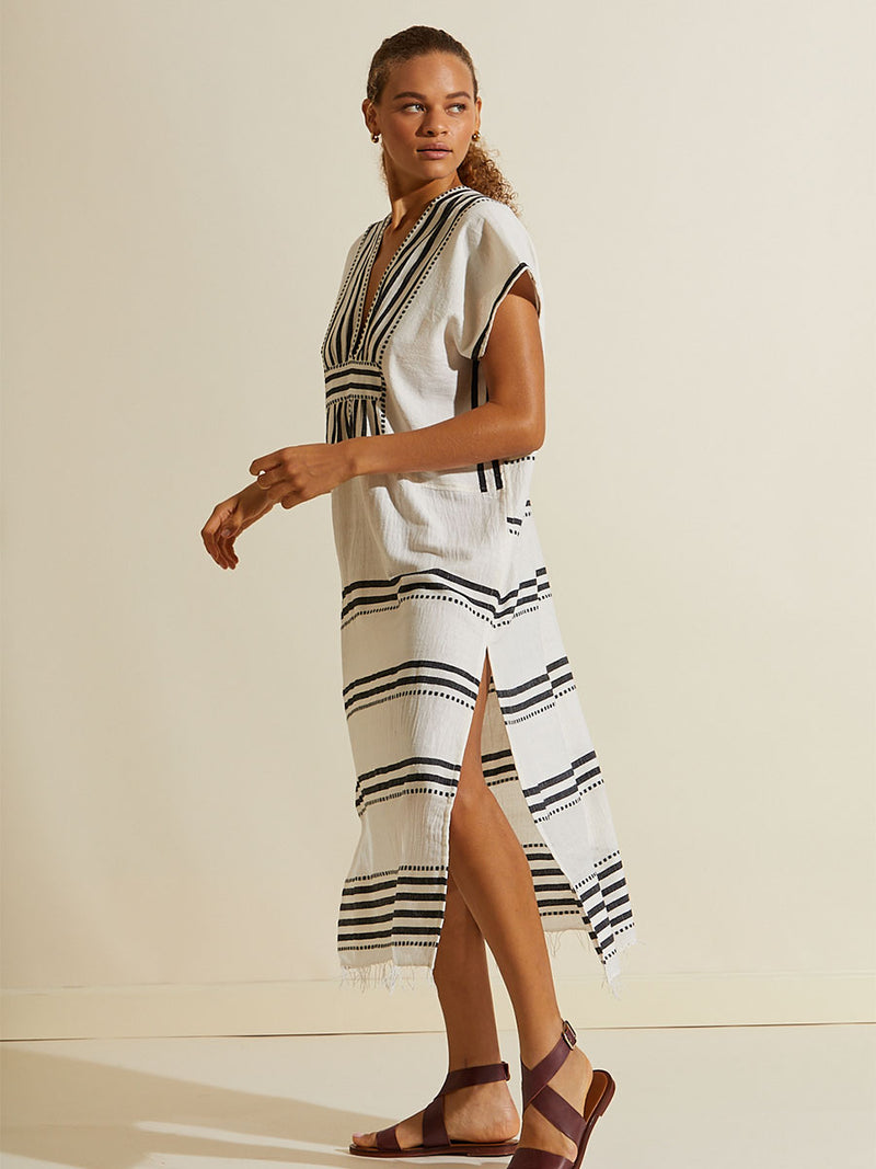 Side view of a woman walking wearing the Eshe Long Caftan Dress featuring architectural and textured black stripes and dotted lines on an off white background.