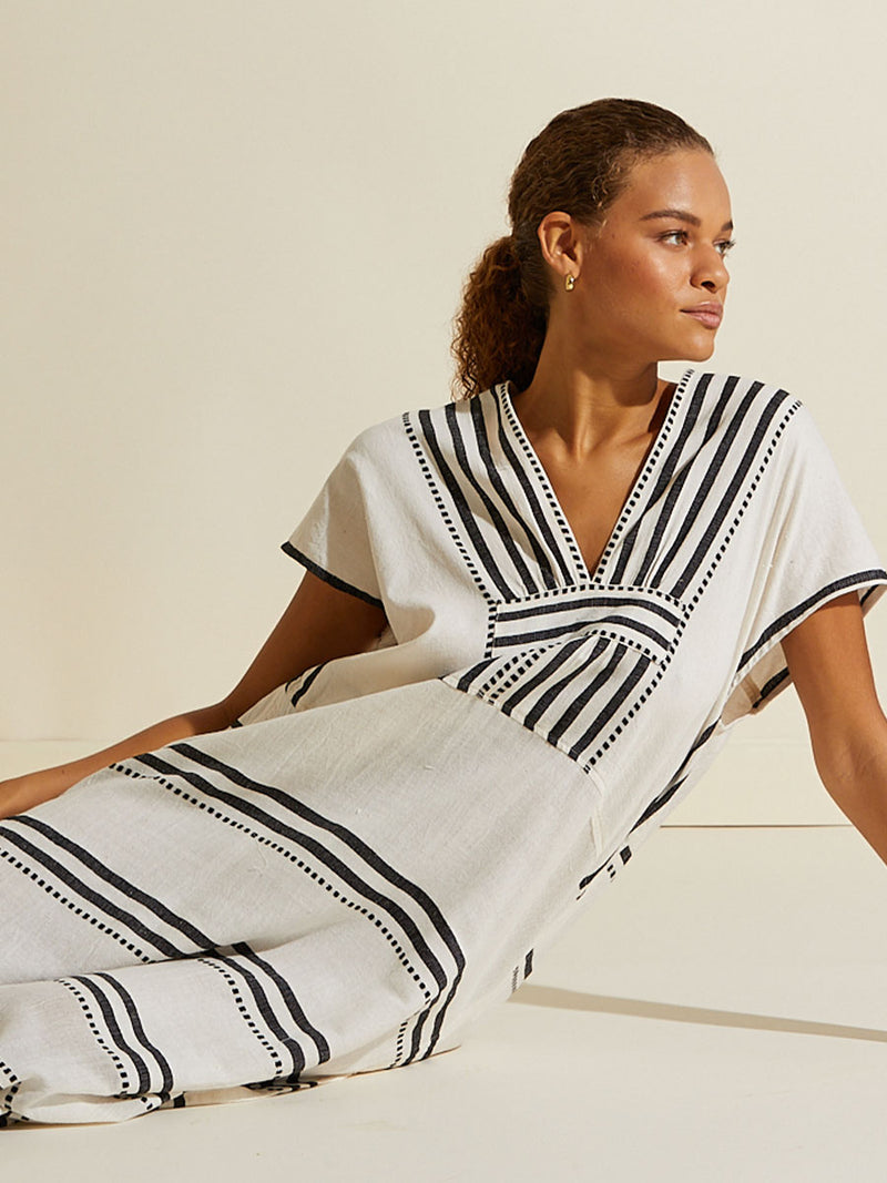 Woman sitting wearing the Eshe Long Caftan Dress featuring architectural and textured black stripes and dotted lines on an off white background.