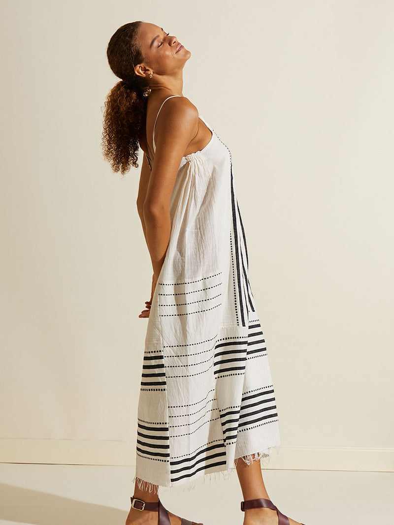 Side view of a woman standing with her hands behind her back wearing the Eshe Slip Dress featuring architectural and textured black stripes and dotted lines on an off white background.