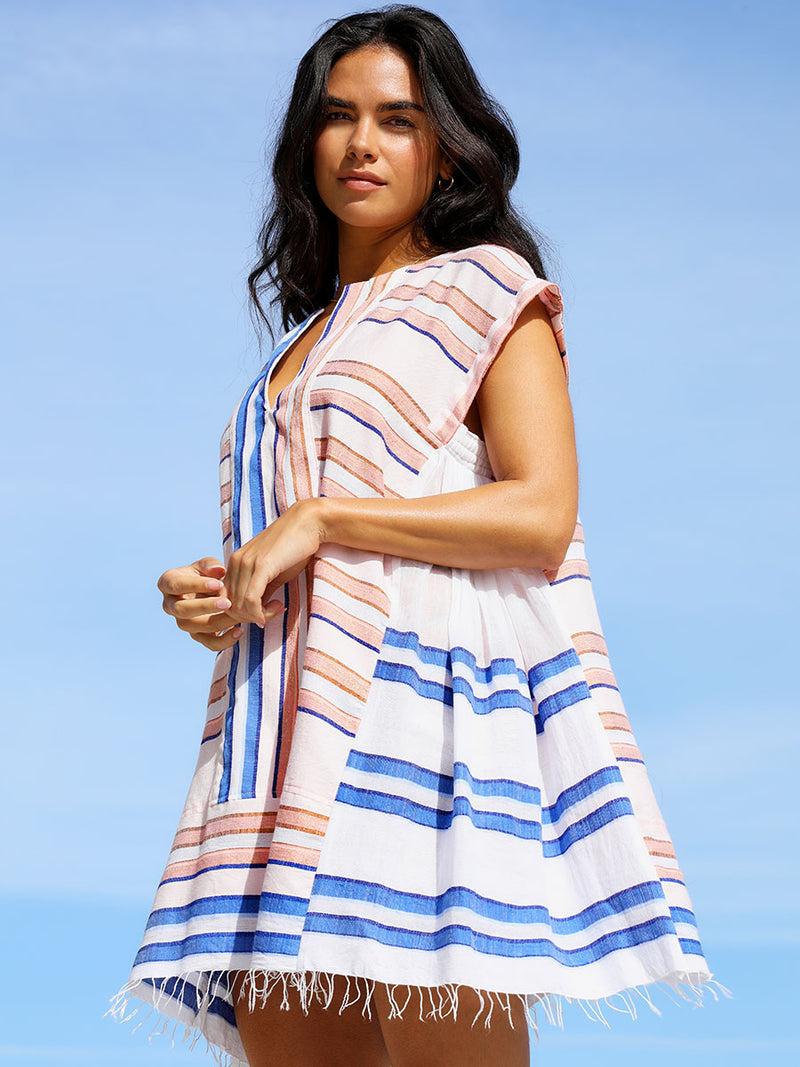 A woman standing wearing the Eskedar Caftan Dress in Seashell featuring blue, white, brown, nude and gold lurex stripes